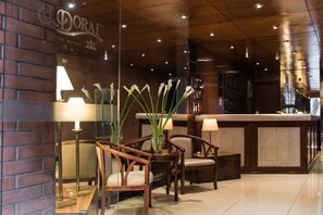 A reception area with a wooden reception desk, two chairs, a vase with white flowers, and a lamp. - El Doral Aparthotel (Lima)