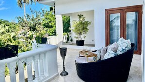 A balcony with a black wicker sofa, a small wooden table with two wine glasses, and a black trash bin. There are potted plants and a palm tree in the background. - Pool Jacuzzi & Tropical Oasis Yard With Fruit Trees (Sosua)