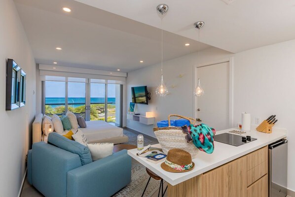 A modern living room with a large window overlooking the sea, a kitchen with a white countertop, and a dining area with a blue sofa.