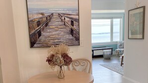 A dining area with a round table, two chairs, a vase with dried flowers, and a large painting of a pier. - The Blue Pearl (Nahant)