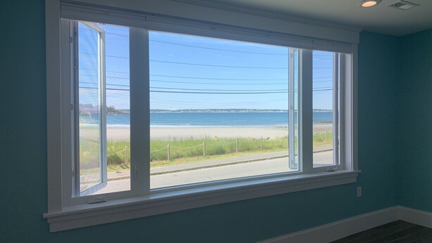 A room with a large window offering a view of a beach and ocean. - The Blue Pearl (Nahant)