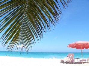 Une plage de sable blanc, avec une eau d’un bleu limpide et un palmier. On y trouve des chaises de plage et un parasol rouge.