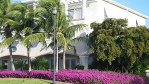 A white building with a balcony, surrounded by palm trees and purple flowers. - Grace Bay Prime Location 2 Bedroom Condo with Beach, Pool and Tennis access!! (Grace Bay)