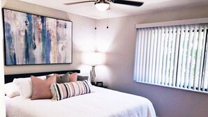 Une chambre avec un grand lit, un ventilateur de plafond, une fenêtre avec des stores et un tableau abstrait accroché au mur.
