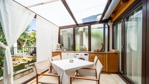 Une terrasse ensoleillée avec une table et des chaises, un lave-linge et un coin cuisine équipé de placards et d’une fenêtre.