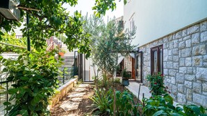 Un étroit sentier de pierre, bordé d’une végétation luxuriante et de plantes en pot, mène à un bâtiment à la façade de pierre et aux portes en bois.