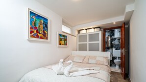 Une chambre à coucher avec un lit, deux tableaux, une salle de bain visible à travers une porte ouverte et un plancher en bois.