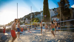 Una cancha de voleibol de playa con arena, con jugadores en movimiento, rodeada por una cerca y edificios al fondo.