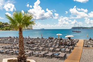 A beachside hotel with rows of sun loungers and a palm tree, a wooden walkway, and a view of the sea with boats. - Apartment & Room Sunset Beach - Double Room (Dubrovnik)
