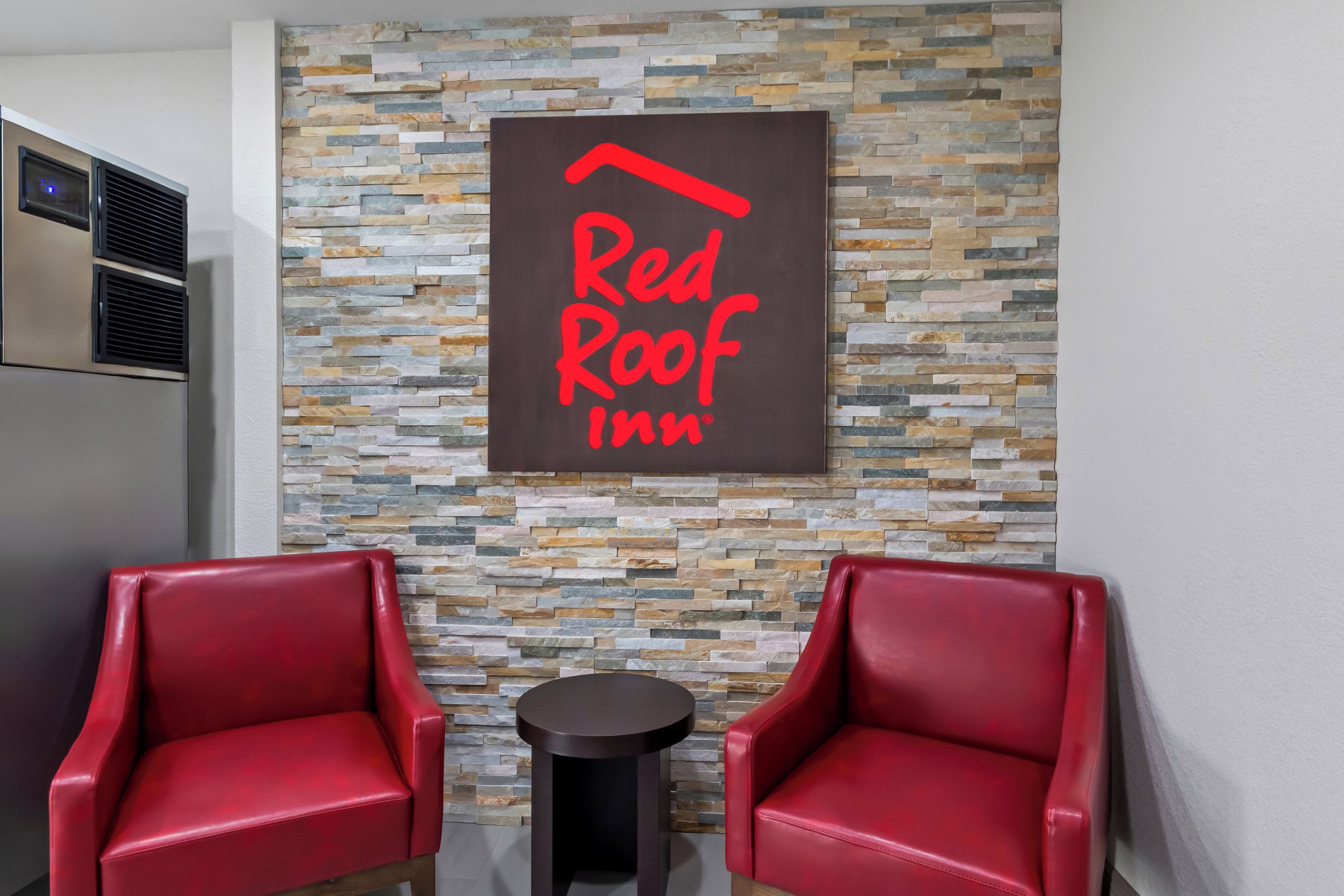 A Red Roof Inn sign on a textured stone wall, two red leather chairs, and a small round table.