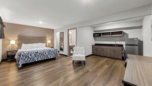 A modern hotel room with a large bed, a small desk, a chair, a kitchenette, and a bathroom.