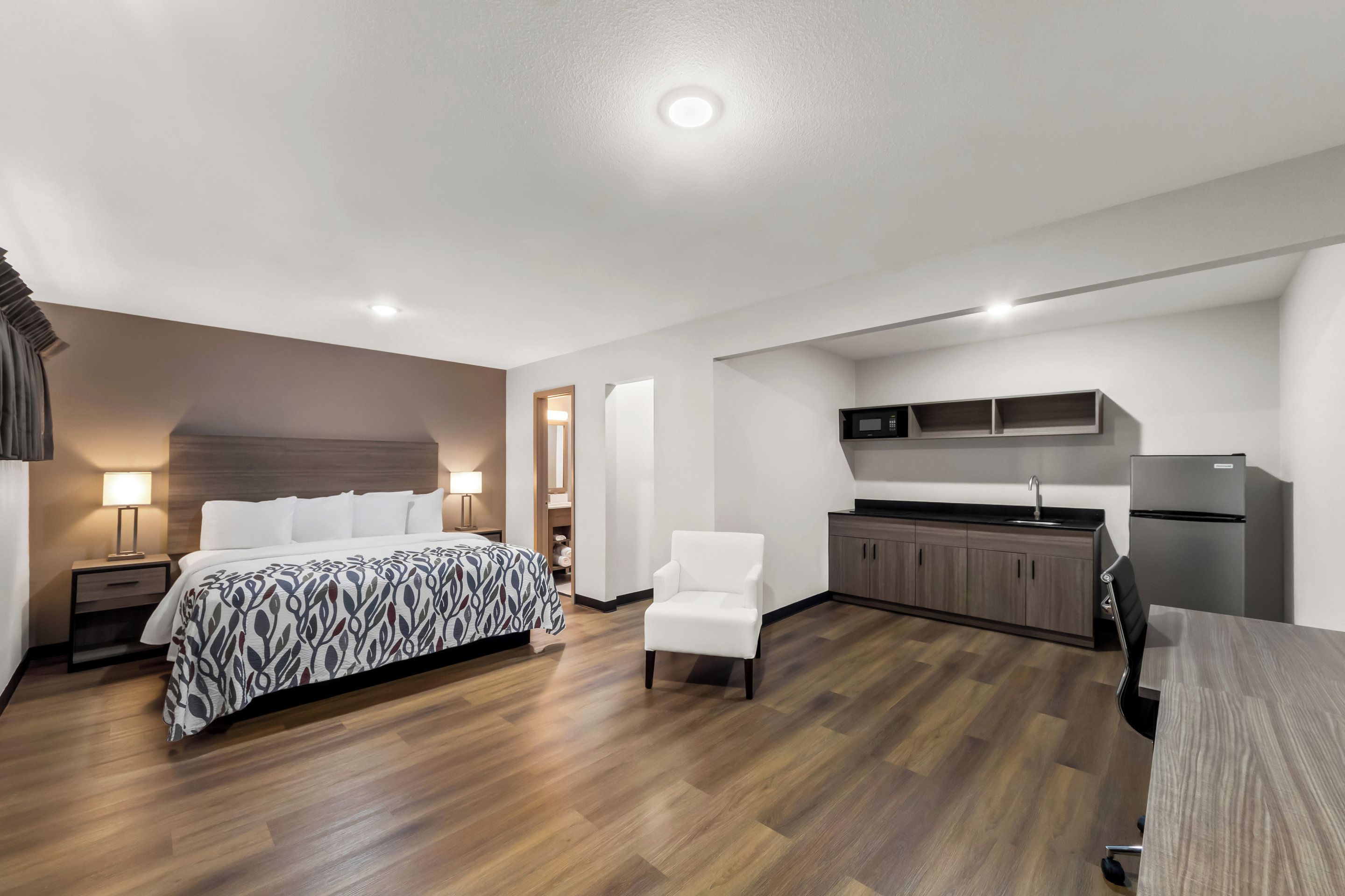 A modern hotel room with a bed, bedside table, lamp, chair, and kitchenette.