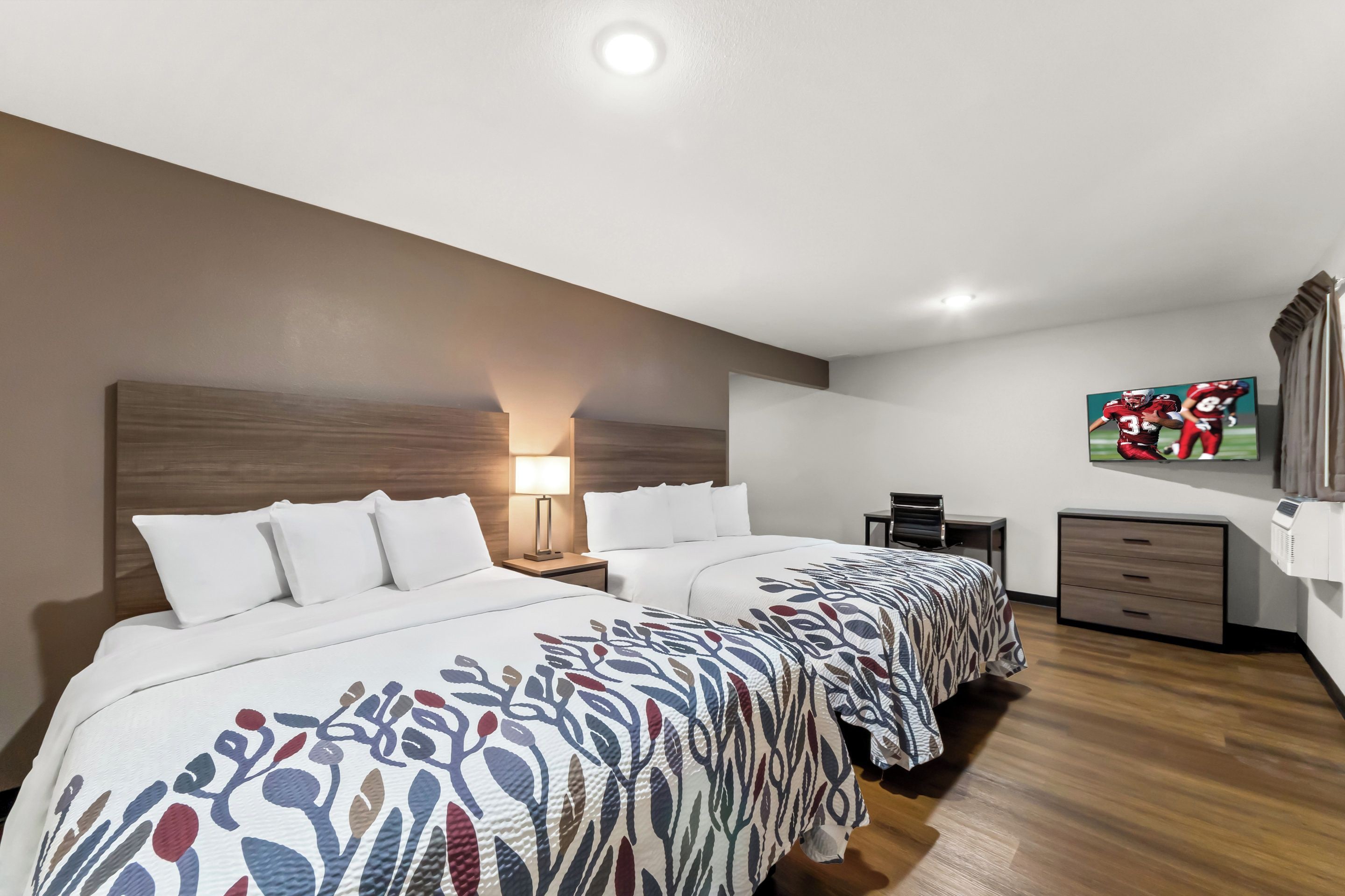 A hotel room with two beds, a nightstand, a desk, a chair, a TV, and a wooden floor.