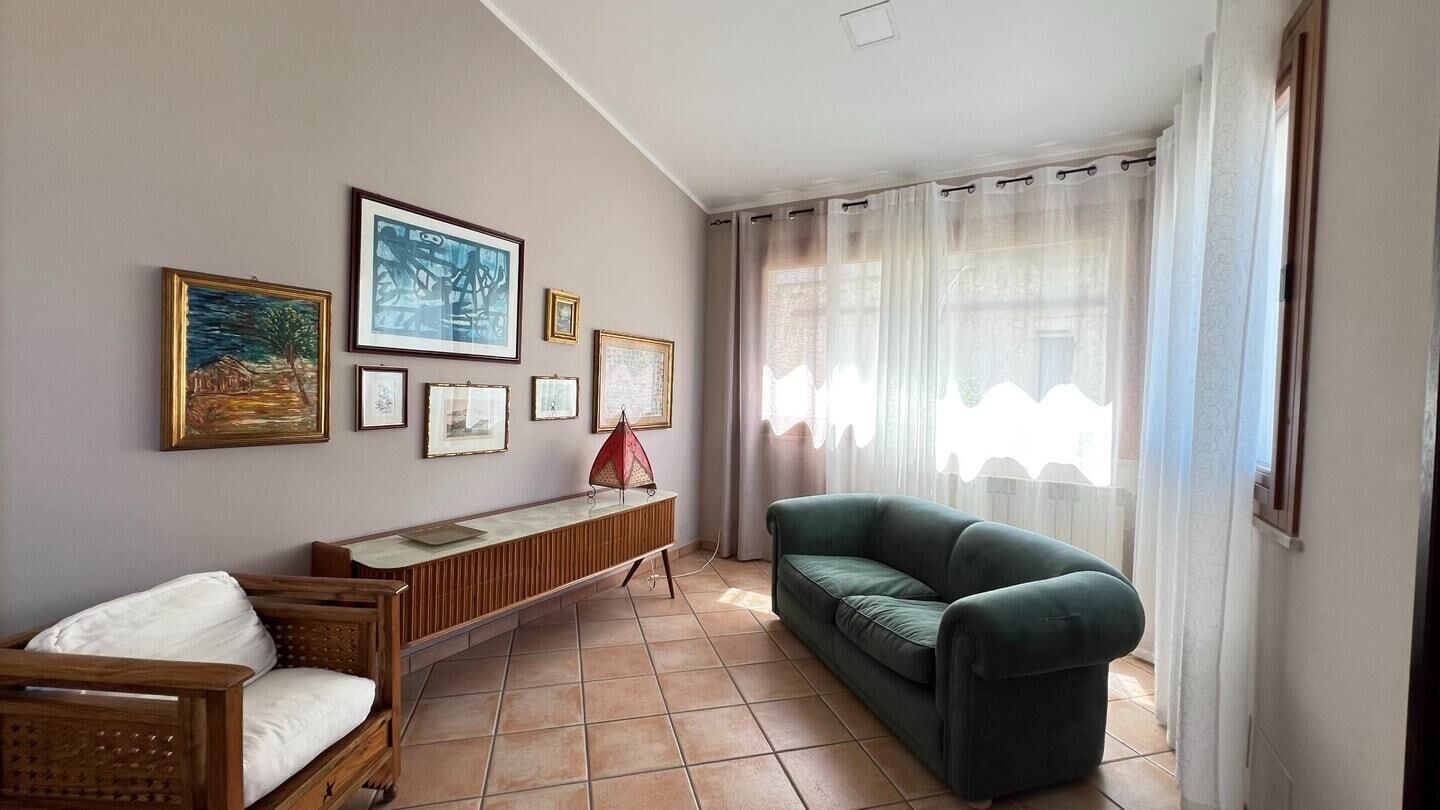 Een woonkamer met een bank, een houten zitbank en een salontafel. Er hangen ingelijste foto’s aan de muur en een raam met gordijnen.
