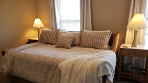 A neatly made bed with a patterned bedspread and pillows, two bedside tables with lamps, and a view of the outdoors through a window with curtains.