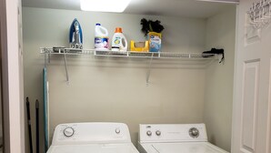A laundry room with two front-loading washing machines, a shelf with cleaning supplies, and a hanging rack.