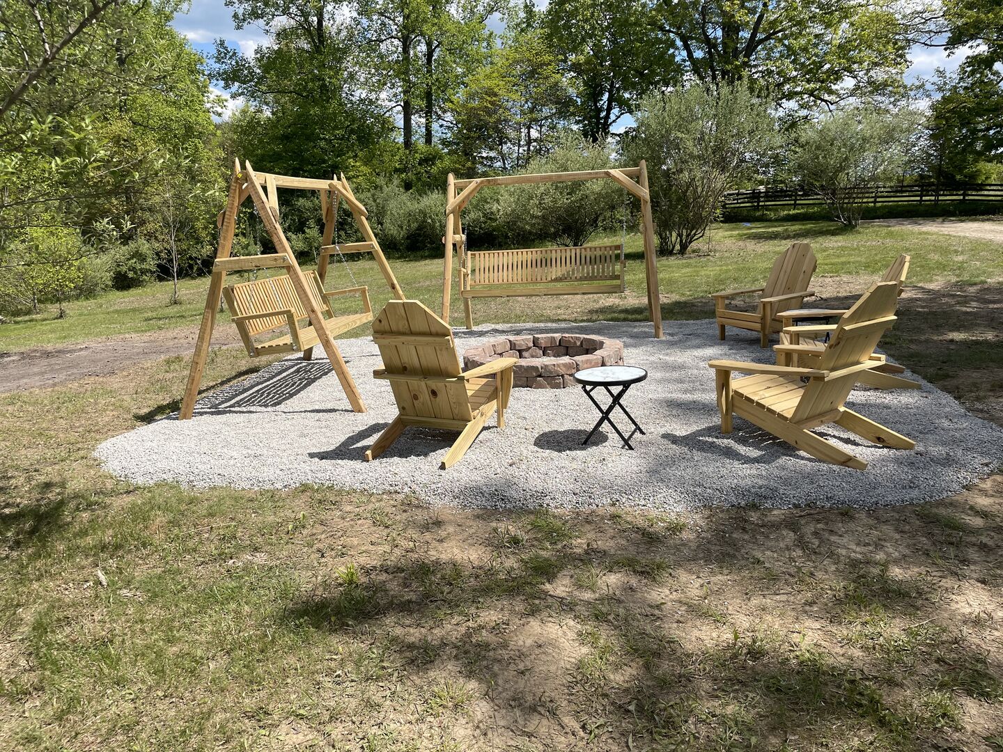 A wooden swing set, Adirondack chairs, and a small round table in a garden area with gravel ground.