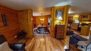 A cabin interior with a wooden dining table, benches, a kitchen area, and a clock on the wall.