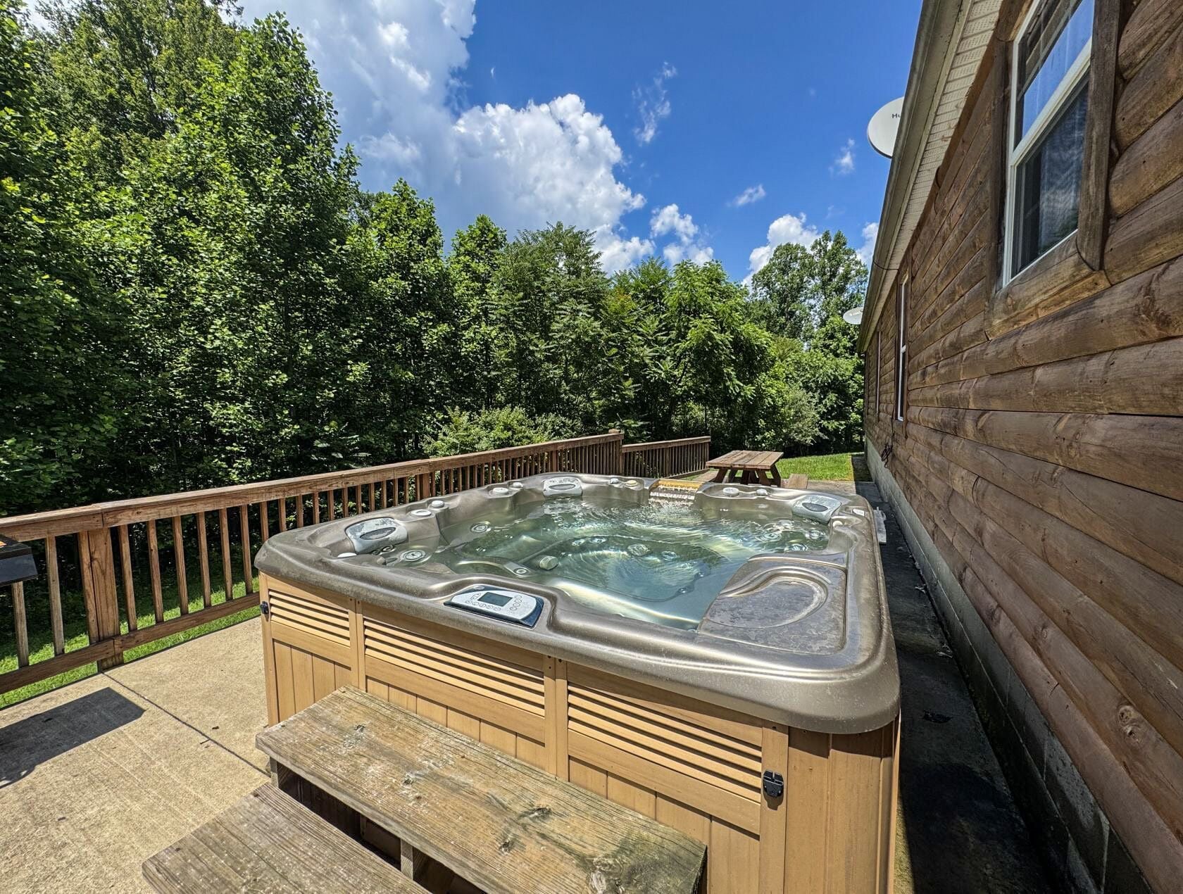 A hot tub with a wooden deck and benches, surrounded by a wooden cabin and lush greenery.