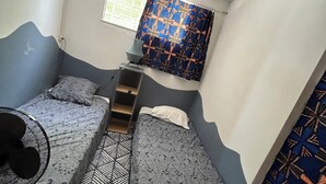 A compact room with two beds, a small table, a fan, and a window with patterned curtains. - Creole-style house with a pool and access to the river (Lamentin)