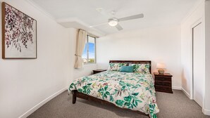 Quarto com cama, mesa de cabeceira, ventilador de teto, janela com cortinas e um quadro na parede.
