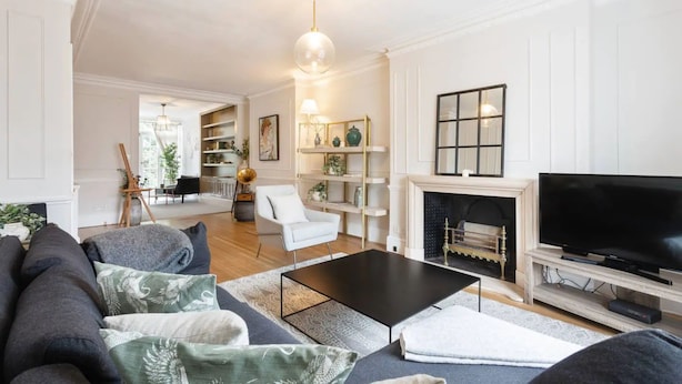 A modern living room with a black coffee table, grey sofa, and white armchairs. There is a fireplace with a TV on a wooden stand. - Beautiful 5 Bedroom Home With Garden in South Kensington (London)