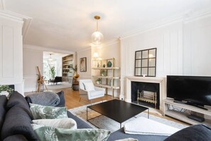 A modern living room with a black coffee table, grey sofa, and white armchairs. There is a fireplace with a TV on a wooden stand.