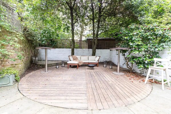 A wooden deck with a sofa and two tables, surrounded by trees and a brick wall.