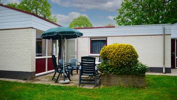 A small, single-story building with a patio area featuring a table and chairs, an umbrella, and a well-maintained hedge.
