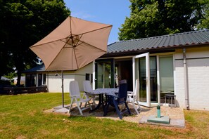 An outdoor patio with a table and chairs, a large umbrella, and a building with a tiled roof and sliding glass doors. - Vakantie Zuid Limburg (Simpelveld)