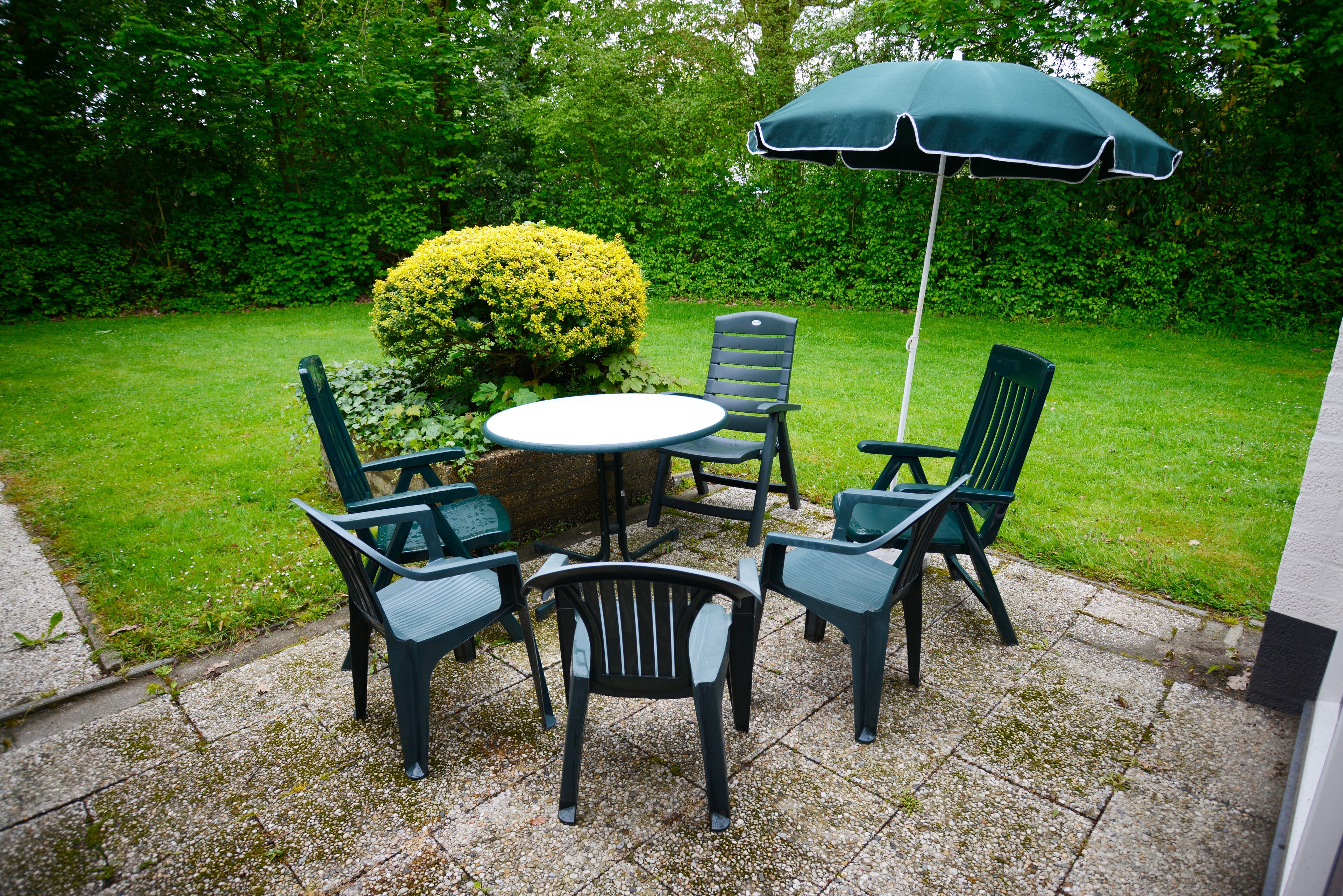 An outdoor patio with a round table and four chairs, a green umbrella, and a well-maintained hedge.