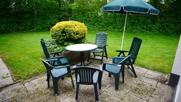 An outdoor patio with a round table and four chairs, a green umbrella, and a well-maintained hedge.