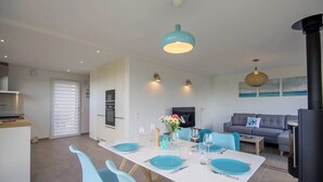 A modern kitchen with white cabinets, a built-in oven, and a dining area with a white table, blue chairs, and teal plates. - Tal Er Forn - PLOUHARNEL (PLOUHARNEL)