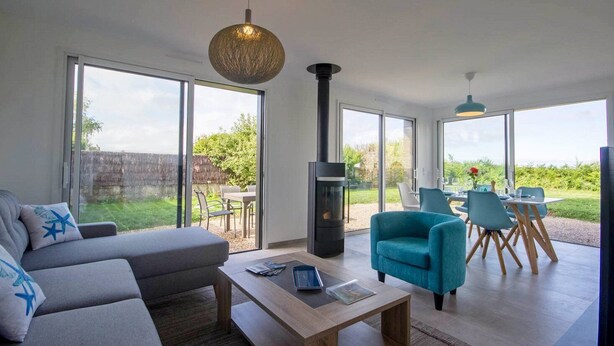 A modern living room with a sofa, a coffee table, and a fireplace. There is a dining area with chairs and a table outside. - Tal Er Forn - PLOUHARNEL (PLOUHARNEL)