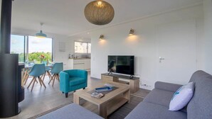 A modern living room with a sofa, a coffee table, and a TV. There is a dining area with chairs and a table, and a kitchen in the background. - Tal Er Forn - PLOUHARNEL (PLOUHARNEL)