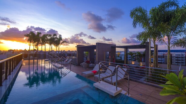 A rooftop pool area with lounge chairs, a small seating area, and a view of the sunset over the city skyline. - Edifício Time - Apto 602 (Maceió)