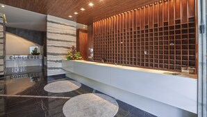 A modern reception area with a wooden panel wall, a marble reception desk, and a glass partition. - Edifício Time - Apto 602 (Maceió)