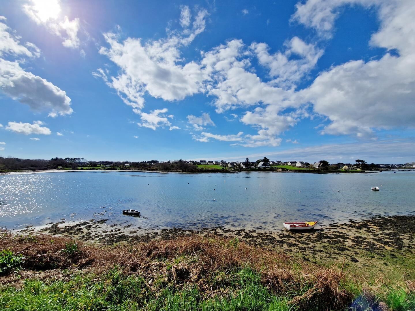 A calm lake with a small boat on the shore, surrounded by grassy areas and a distant village.