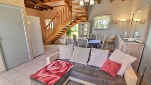 A cozy living room with a sofa, a dining table, and a staircase leading to another room. - L'Hortensia - PLEUDANIEL (PLEUDANIEL)