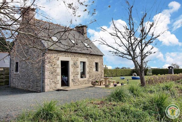 Une petite maison en pierre avec une porte ouverte, un petit jardin avec des plantes en pot et une haie en arrière-plan.