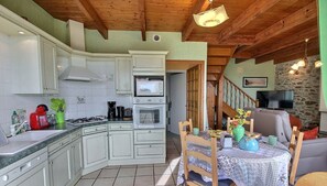 Una cocina con armarios blancos, microondas y estufa. Hay una mesa de comedor con sillas y un televisor al fondo.