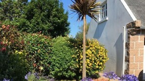 A garden with various plants and flowers, a palm tree, and a building with a sloped roof. - Villa Sérénité - PERROS GUIREC (PERROS GUIREC)