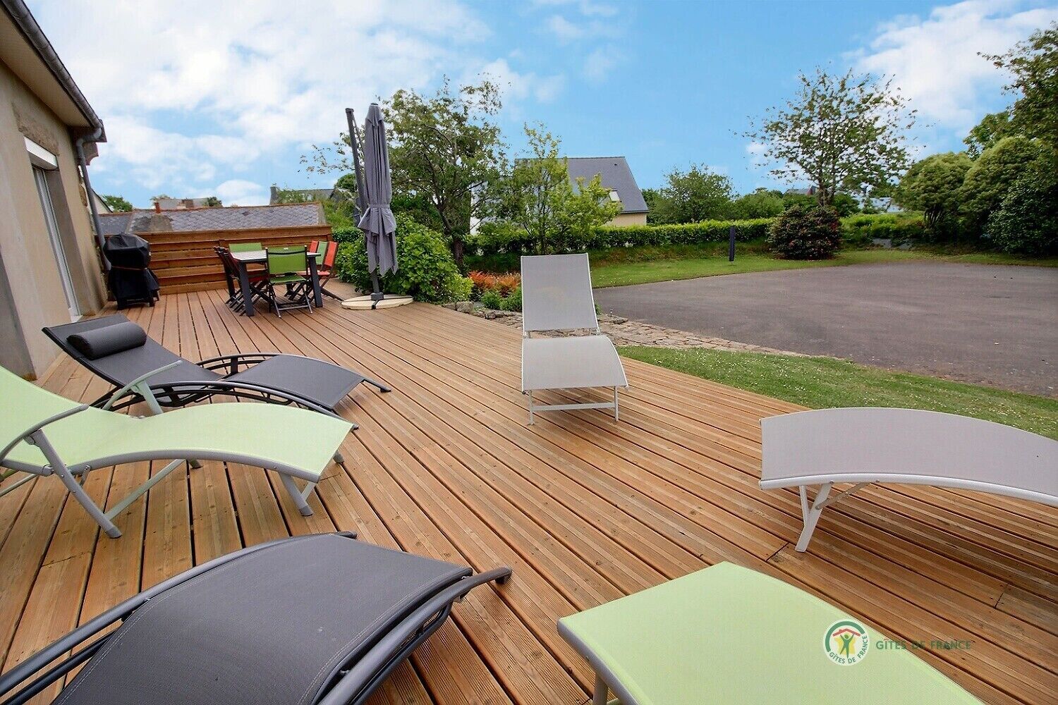Eine Holzterrasse mit Gartenmöbeln, darunter Liegestühle und ein Tisch, mit Blick auf einen Garten und eine Straße.
