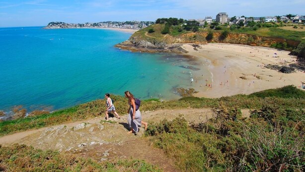 A coastal landscape with a sandy beach, clear blue sea, and a path leading to the shore.
