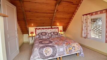 A bedroom with a sloped ceiling, a bed with patterned bedding, a bedside lamp, and a window with curtains.