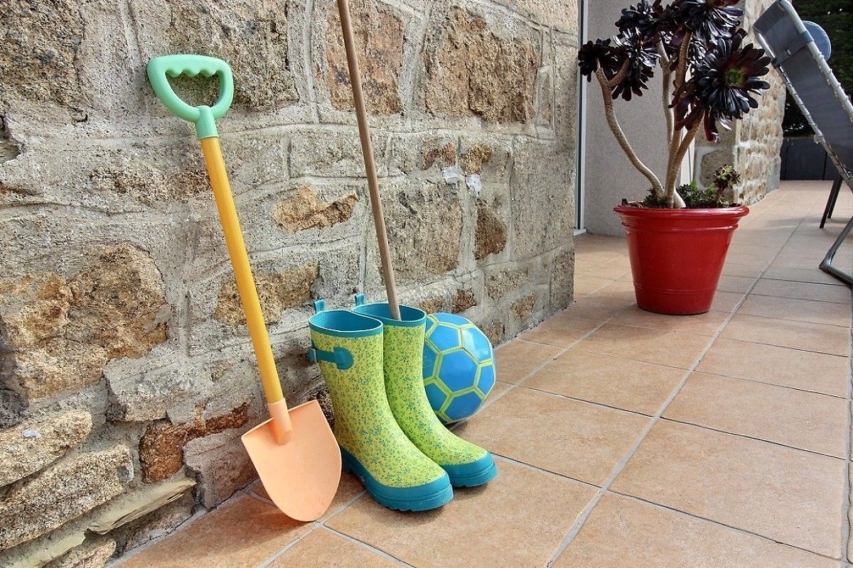 A pair of green rain boots, a shovel, and a soccer ball leaning against a stone wall.