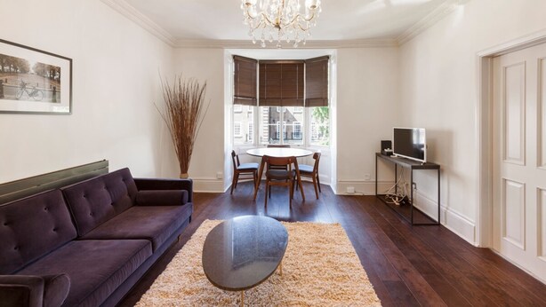 A living room with a dark-colored sofa, a round coffee table, a chandelier, a dining table with chairs, and a television on a stand. - Elegant, Airy 2 Bedroom Flat in Paddington (London)