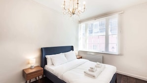 A bedroom with a large bed, a chandelier, a nightstand with a lamp, and a window with blinds. - Elegant, Airy 2 Bedroom Flat in Paddington (London)