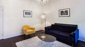A modern living room with a dark blue sofa, a yellow armchair, a round coffee table, and a floor lamp. There are two framed pictures on the walls. - Elegant, Airy 2 Bedroom Flat in Paddington (London)