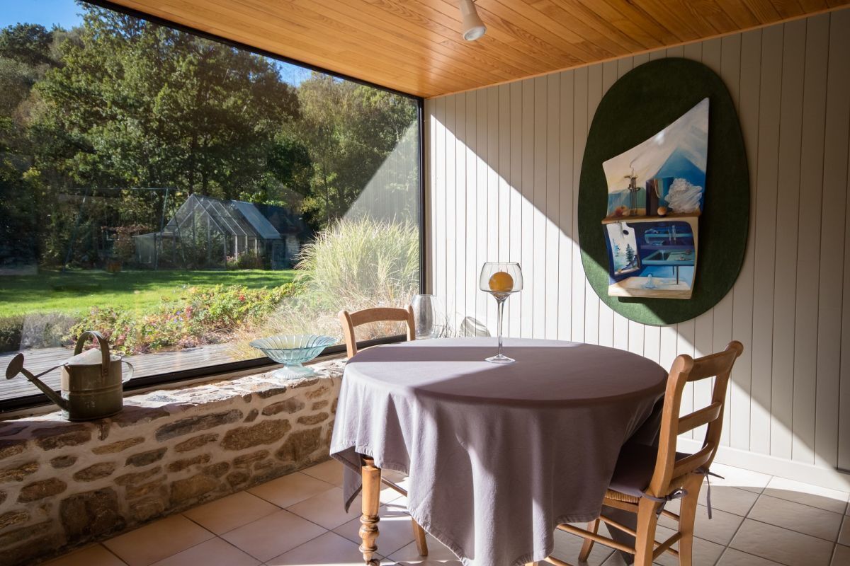 A dining area with a table and chairs, a painting on the wall, and a view of a greenhouse and garden.